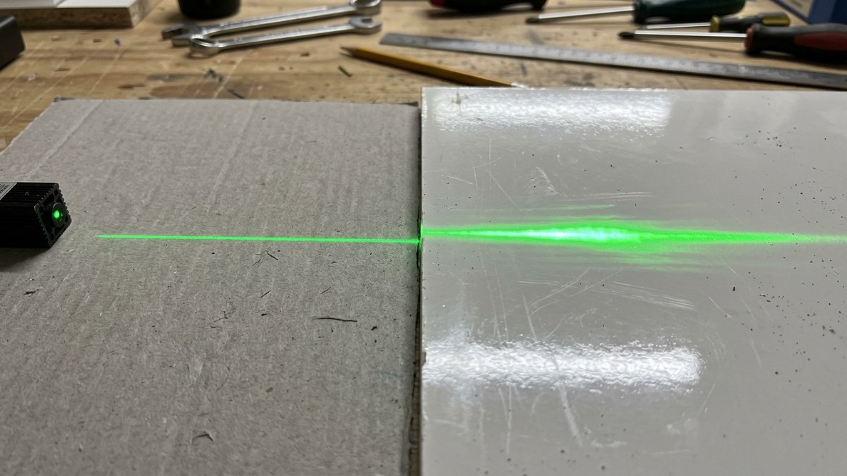 Groene laserlijn valt over een mat stuk karton en een glanzend werkblad, waarbij de lijn op het matte deel duidelijk scherper is